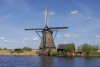 A windmill and a shed next to a body of water, surrounded by reeds under a sunny blue sky, many