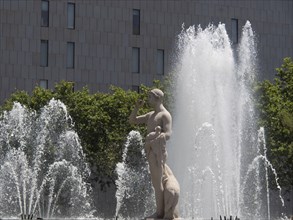 A fountain with a statue surrounded by water fountains and trees in daylight in front of a modern