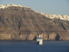A large cruise ship sails along the coast of Santorini, past steep cliffs and white buildings,