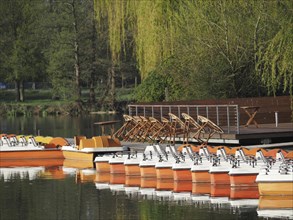 Several pedal boats in orange and white on the jetty of a lake surrounded by green vegetation,