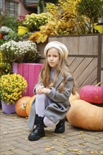 Little girl in a beret and wool coat sits on squats next to colorful pumpkins