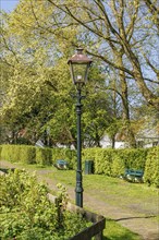 An antique lamppost stands in a well-kept park, surrounded by trees and green bushes on a sunny