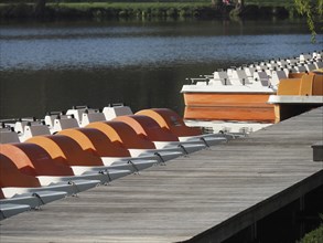View of a jetty with several orange and white pedal boats on a calm lake, rowing boats and pedal