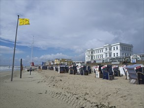 View of beach chairs on the beach in front of buildings and a yellow flag under a cloudy sky, blue