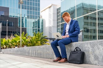 Horizontal full length photo of a businessman using laptop sitting outside a financial building