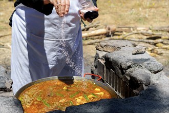 Woman cook in white apron adding salt to a Spanish paella cooked in the countryside