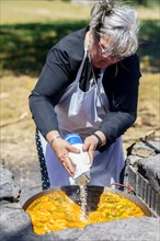 Woman cook in white apron pouring rice into a typical Spanish paella cooked in the countryside on