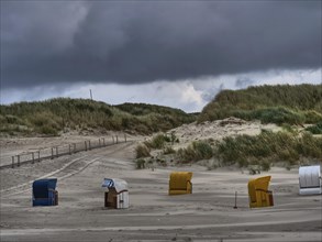 Path through dunes to the beach with dark clouds, scattered beach chairs, colourful beach chairs on
