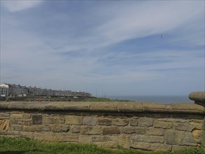 Stone wall with a view of houses and the sea under a cloudy sky, green surroundings, ruins and old