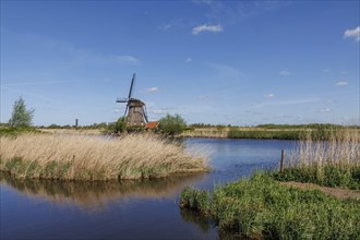Windmill stands on the river bank, surrounded by reeds and grass under a clear blue sky, many