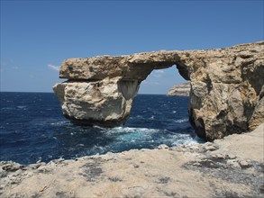 Rock arch over the sea, surrounded by cliffs, under a clear blue sky, many historic windmills by a
