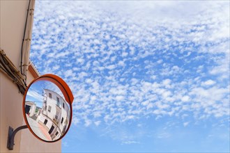 Traffic mirror on the facade of a building reflecting a street with a cloudy sky