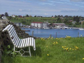 White bench on a green meadow with a view of the sea, yachts and a small village in the background,