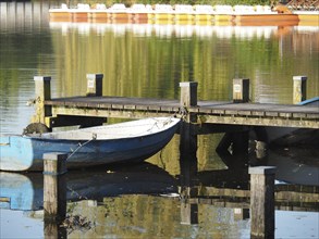 A boat is moored next to a jetty that is reflected in the water, surrounded by peaceful nature,