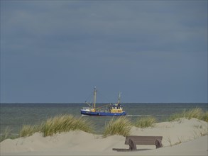 A fishing boat on the sea, in the foreground dunes and a bench under a cloudy sky, dunes on an