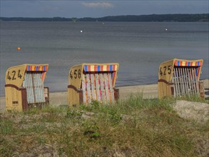 Three beach chairs with colourful stripes stand on a sandy beach in front of a calm sea and cloudy