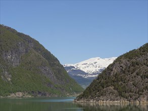 A narrow lake between large rocky mountains, with snow-capped peaks in the background and a blue