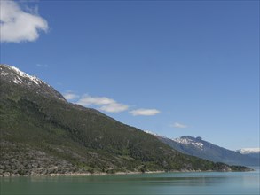 Landscape shot of a lake and forested mountains under a clear blue sky, greenish shimmering water