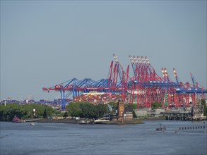 Container cranes at the harbour with a river and buildings in the foreground, under a blue sky and
