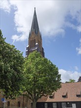 A church tower rises behind a large tree in a village with brick houses and cloudy sky, large