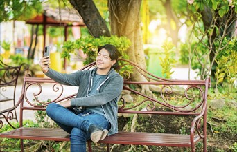Smiling man sitting on a bench taking a selfie in Nagarote Park. Relaxed guy taking a selfie