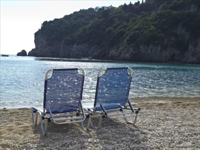 Two blue deckchairs on the beach with calm seascape and rocks in the background, umbrellas and