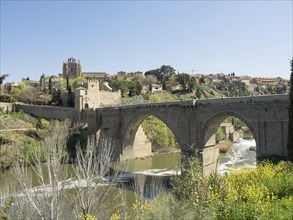 Historic stone bridge with arches over a river, surrounded by vegetation and cityscape, sunny