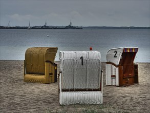 Three beach chairs stand on the sandy beach in front of a calm sea under a cloudy sky. Ships are