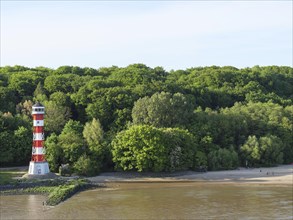Red and white lighthouse on the river bank, surrounded by lush greenery and trees under a clear