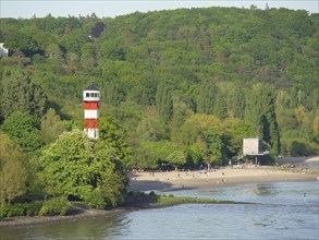 People on the beach near a red and white lighthouse, surrounded by dense forests, green shore on a