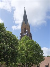 A tall church tower with a green roof rises into the overcast sky between green trees, large church