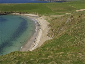 Steep cliffs and a narrow sandy beach surrounded by green grassland and blue water under a clear