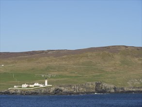 A lighthouse and several buildings stand on a rocky coastline against grassy hills and a clear blue