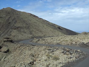 A winding road meanders through a barren, stony volcanic landscape under a blue sky, barren
