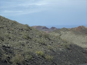 Hills of a volcanic landscape with sparse vegetation and stony slopes under a blue sky, barren