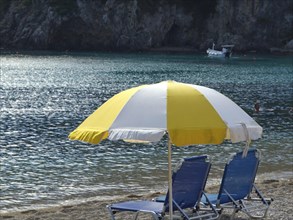 Yellow and white parasol and two deckchairs on the beach with sea and coast in the background,