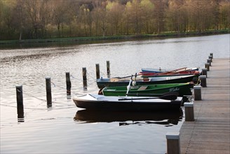 Several small boats are moored at a pier on calm lake water, surrounded by spring trees, rowing
