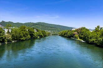 A calm river flows through a landscape of green trees and hills under a blue sky, Olten, Solothurn,