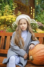 Cute little girl in a beret and woolen coat sits on bench leaning on pumpkin