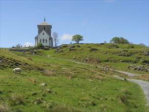 Church on hilly landscape with hiking trail and sheep, blue sky and clouds, old stone church and