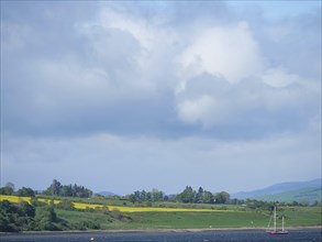 Landscape with hills and fields seen across the water under a cloudy sky, blooming yellow fields on
