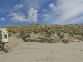View of a grassy sand dune with a small raised house and a blue sky, clouds on the beach with dunes