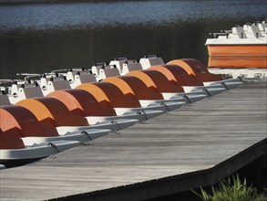 Close-up of a jetty with several orange and white pedal boats along a calm lake, rowing boats and
