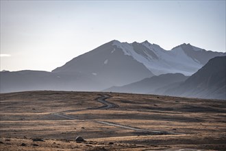 Gravel road through autumnal plateau with brown grass, glaciated and snow-covered peaks, Ak Shyrak