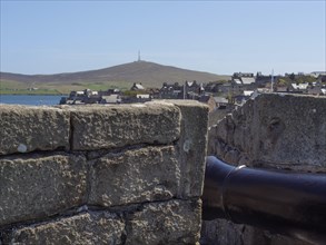 A cannon looking over a stone wall at a town with a hill in the background, old cannons on a stone