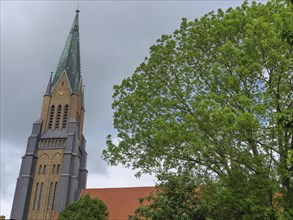 A tall church tower behind green trees rising into the cloudy sky, large church towers with trees