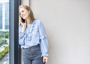 A beautiful mid-adult blonde woman is talking on the phone while she looks through the window
