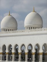 Mosque with two large white domes and golden spires, surrounded by arches, under a blue sky,