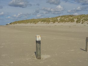 Sandy coastal area with grass-covered dunes and a post in the foreground, clouds on the beach with