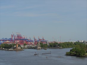 Container cranes on the harbour shore with water, trees and buildings in the foreground under a